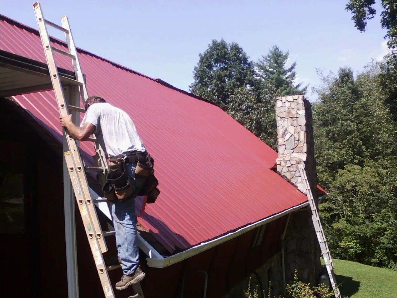 Roofer on a ladder beside a red metal roof and stone chimney — Soffit & Fascia Repair by Roofing Brothers in Cannon Beach